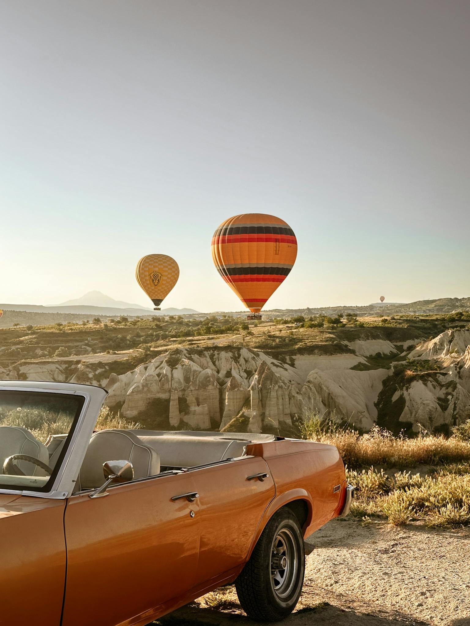Classic car with colorful balloons flying over Cappadocia's unique landscape, captured at sunrise.